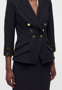 Gros plan d’un blazer noir à double boutonnage avec des boutons dorés et des détails aux poignets, porté sur une jupe noire.
