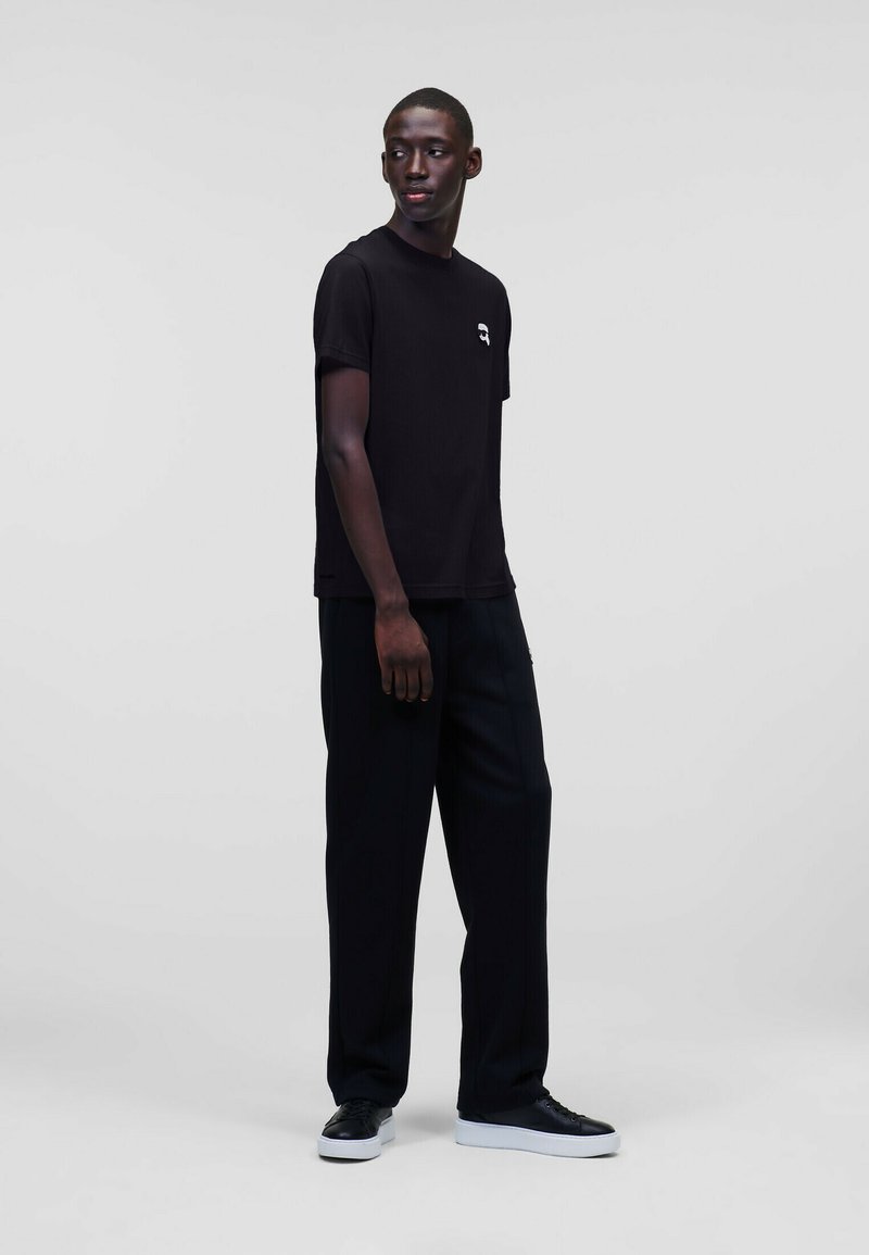 T-shirt nera in cotone con scollatura rotonda, maniche corte e piccolo grafico bianco sul petto, abbinata a pantaloni neri larghi e sneakers.