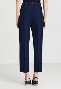 Dunkelblaue, hoch taillierte Hose mit einem dehnbaren Bund, geraden Beinläufen und zwei Gesäßtaschen. Getragen mit schwarzen Absätzen.