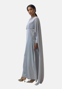 Robe longue et fluide en satin gris clair. Présente un design en couches, des manches longues et un dos en forme de cape. Portée avec des talons beiges.