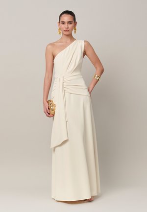 Femme portant une élégante robe crème asymétrique à une épaule avec drapés, boucles d'oreilles dorées imposantes, bracelet manchette, et tenant une pochette dorée.
