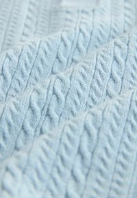 Helles blaues Textil mit einer Kabelstrickmuster, das erhabene, ineinander verwobene Designs in weichen, gleichmäßigen Schlaufen präsentiert.
