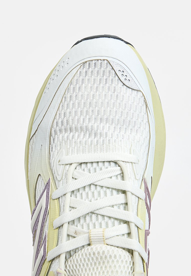 Zapatilla deportiva ligera en blanco y verde pálido, con una parte superior de malla texturizada, suela gruesa y acentos morados a los lados.