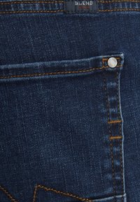Mörkblå denimjeans med en bakficka med orange sömnad och en silverfärgad knappdetalj. Tyget har en slät struktur.