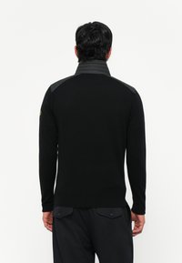 Άνδρ�ας ντυμένος με μαύρο μπουφάν μακρυμάνικο με επενδεδυμένους ώμους και γιακά, στέκεται στραμμένος μακριά μπροστά σε λευκό φόντο.