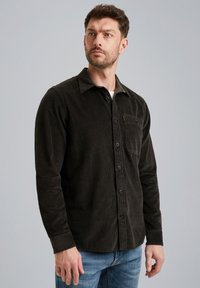 Camisa de pana oscura con botones y un bolsillo en el pecho, con un cuello sólido y mangas largas, llevada sobre una camiseta blanca y unos vaqueros azules.