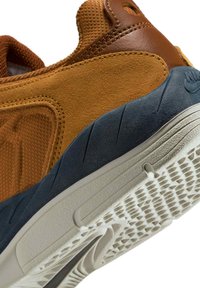 Brązowo-beżowe sneakersy z zamszu z teksturowaną siateczką, niebieską środkową częścią podeszwy i białą wzorzystą podeszwą, ustawione na tle białym pod kątem.