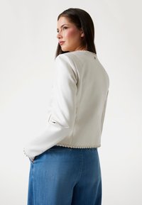 Witte neplederen jas met een cropped ontwerp, gedetailleerde stiksels langs de zoom en een ronde halslijn. Gecombineerd met blauwe denim.