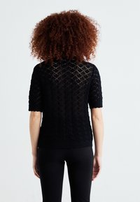Pull en tricot noir à manches courtes, avec un motif en losanges et un ourlet à côtes. Le dos présente un élégant travail de dentelle.