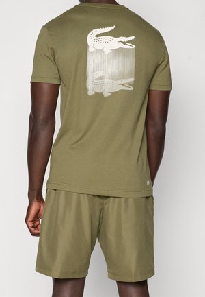 Mand iført olivengrønne shorts og T-shirt med et hvidt motiv af en krokodille og dens spejlbillede på ryggen.