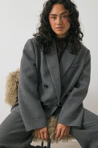 Sivi dvoredni kostim iz teksturirane tkanine z velikimi črnimi gumbi, širokimi reverji in sproščenim krojem. Model sedi na puhasnem stolu.