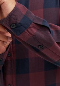 Camicia a quadri con colori bordeaux e blu navy, polsini abbottonati, materiale in cotone morbido e finitura testurizzata con dettagli di cucitura visibili.