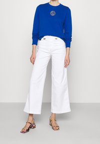 Blauwe sweater met lange mouwen en logo, gecombineerd met witte, wijde, hoog getailleerde broek en sandalen met blokhak en bloemmotief.