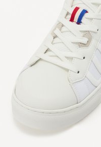 Fehér sneaker sima bőr felsőrésszel, hálós díszítésekkel és ellentétes kék és piros csíkos részletekkel a nyelvén. Lapos cipőfűző, textúrált talp.
