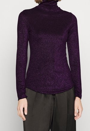 Nainen pukeutunut istuvaan, pitkähihnaiseen, hohtavaan purppuranväriseen poolopaitaan ja väljissä tummissa housuissa, yksinkertaista taustaa vasten.