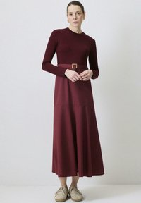 Maroon långärmad klänning med figurnära överdel och utsvängd kjol, försedd med ett bälte med en guldbuckles accent. Slät, mjuk tygtextur.