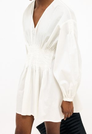 Personne portant une robe blanche avec une taille froncée et de longues manches bouffantes, tenant une pochette tissée noire.