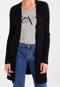 Schwarze gerippte Strickjacke mit offenem Vorderteil, langen Ärmeln und zwei Fronttaschen, getragen über einem grauen Grafik-T-Shirt und blauen hochgeschnittenen Jeans.