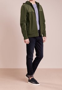 Mørkegrøn hættetrøje med lynlås og en kontrasterende øverste del, kombineret med mørke jeans og sorte sneakers. Minimalistisk design med en tekstureret overflade.