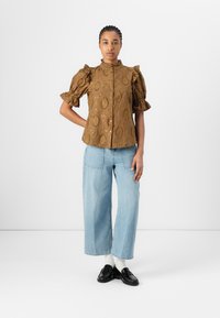Blusa marrone con maniche a palloncino, motif paisley testurizzato, chiusura anteriore a bottoni e colletto arricciato. Abbinata a jeans a gamba larga di colore blu chiaro e mocassini neri.