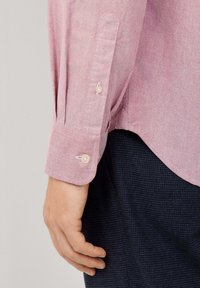 La mano de un hombre al lado de una camisa de manga larga rosa con puños y dos botones, combinada con pantalones oscuros de textura azul.