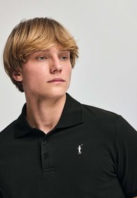 Svart polotröja med en texturerad tyg, med en knäppning i två knappar och en liten vit golfarelogotyp på vänster bröst.