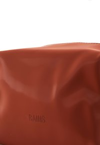 Bolsa impermeable roja con una textura suave y brillante y una apariencia ligeramente arrugada, que cuenta con la palabra "RAINS" en relieve en la parte frontal.