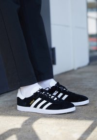 Melnas zamšādas sporta kurpes ar baltām akcentiem un trim svītrām, ar baltu zoli un zelta "Gazelle" uzrakstu sānos.