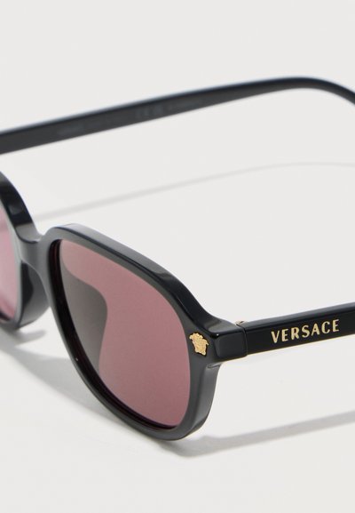 Μαύρα ορθογώνια γυαλιά ηλίου Versace με σκούρους μωβ φακούς και χρυσό λογότυπο Μέδουσας στη γωνία του σκελετού και το όνομα της μάρκας στον βραχίονα.