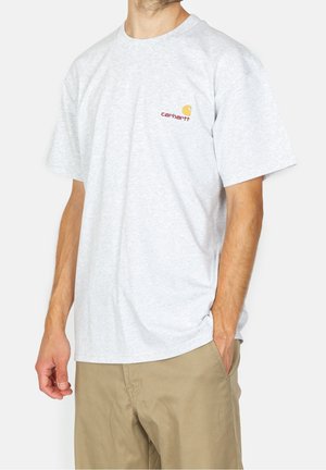 Mann trägt ein hellgraues Carhartt-T-Shirt und beige Hose, mit einer Hand in der Tasche, vor einem schlichten weißen Hintergrund.