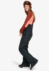 Conjunto de esquí que incluye una parte superior en tonos bloqueados de marrón rojizo y rosa con un mono negro y botas negras; incluye gafas de esquí y un gorro de punto.