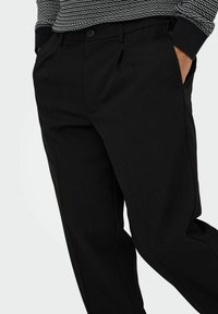 Schwarze, maßgeschneiderte Hose aus glattem Material, mit flachem Frontschnitt, Seitentaschen und zwei Knöpfen am Bund zur Befestigung.