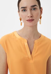 Oranges ärmelloses Top mit V-Ausschnitt. Glatte Stoffoberfläche mit minimaler Textur. Große goldene Ohrringe ergänzen das Outfit.