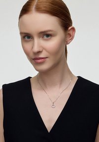 Srebrny naszyjnik z okrągłym wisiorkiem, ozdobiony małymi kamieniami dekoracyjnymi. Modelka nosi czarną bluzkę oraz minimalistyczne kolczyki.