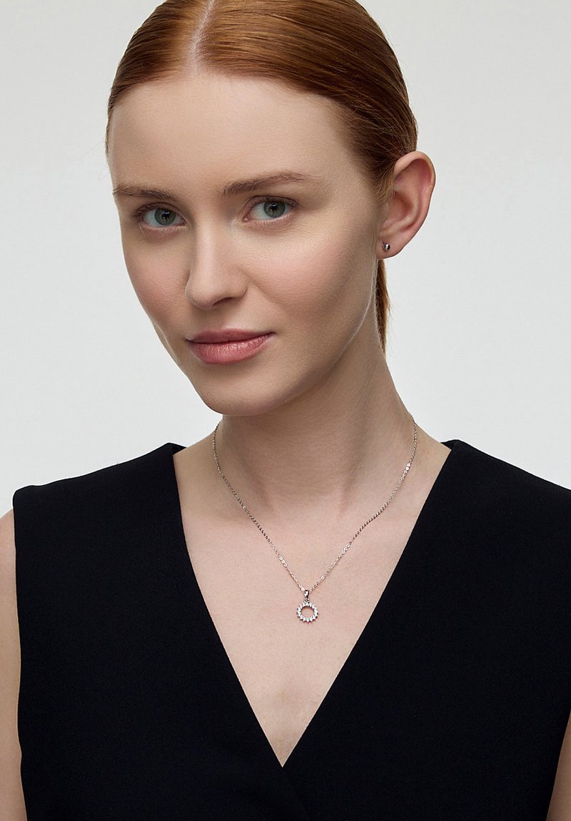 Srebrny naszyjnik z okrągłym wisiorkiem, ozdobiony małymi kamieniami dekoracyjnymi. Modelka nosi czarną bluzkę oraz minimalistyczne kolczyki.