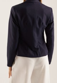 Blazer azul marino con un diseño ajustado, textura suave y hombros estructurados. Cuenta con una sola abertura en la parte trasera y mangas largas, combinado con pantalones blancos.