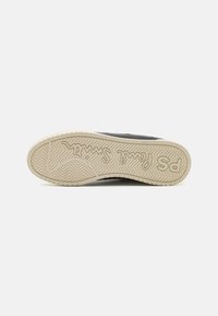 Zapatilla negra con una suela de goma con un patrón de espiga y un logotipo en relieve. La entresuela es de color crema con una textura detallada.