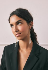 Zlatne uhane z obeski, s tankimi, teksturiranimi verigami, visijo pod ušesno mečico in dopolnjujejo črn blazer ter nevtralno ozadje.