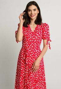 Czerwona sukienka w kwiaty zapinana na kokardę w talii z krótkimi bufkami. Posiada biały wzór kwiatowy i dekolt w serek. Gładka tekstura materiału.