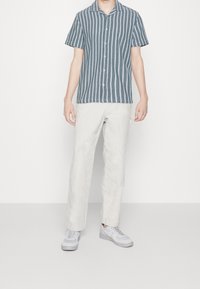 Košile s krátkým rukávem s modro-bílými vertikálními pruhy, spárovaná se světlými kalhotami a šedými teniskami. Materiál vypadá lehce a texturovaně.