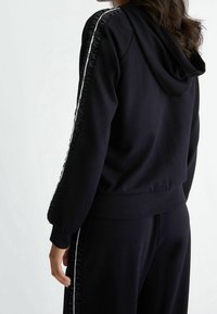Zwarte hoodie van zachte stof, met een capuchon, ruches aan de zijkanten en witte accentstrepen langs de mouwen.