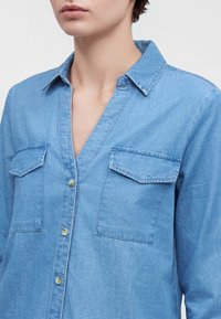 Gros plan d'une personne portant une chemise en denim bleu clair avec fermeture à boutons et deux poches poitrine avant avec rabats.