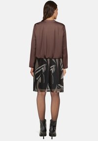 Brązowa satynowa bluzka z długim rękawem, zestawiona z przezroczystą spódnicą w czarny kwiatowy wzór; w połączeniu z czarnymi botkami za kostkę, uwydatnia warstwowy design.