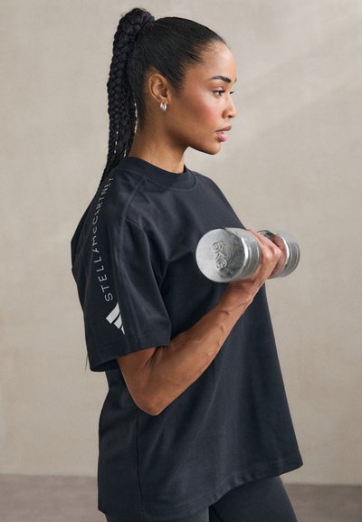 T-shirt noir surdimensionné avec détail de logo sur la manche, porté en tenant un haltère en métal. Le design simple et la texture douce sont mis en avant.