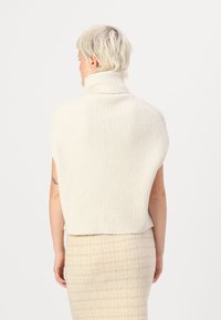 Kremasta pletena turtleneck pulover s rebrasto teksturo, kratkimi rokavi in sproščenim krojem. Nosi se čez bež textured krilo.