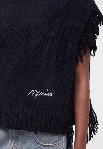 Pull sans manches en tricot noir avec bords à franges et logo Moschino blanc au-dessus de la poche d'un jean bleu clair.
