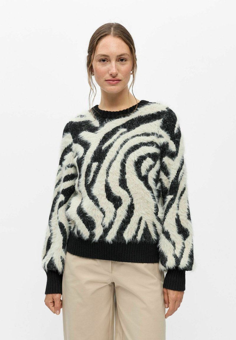 Femme à la peau claire et aux cheveux bruns portant un pull duveteux à rayures zébrées noires et blanches et un pantalon beige sur un fond blanc uni.
