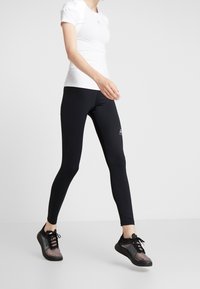 Černé legíny a upnuté bílé tričko jsou doplněny černými sportovními botami. Oblečení je navrženo pro aktivní použití se štíhlým, uhlazeným vzhledem.