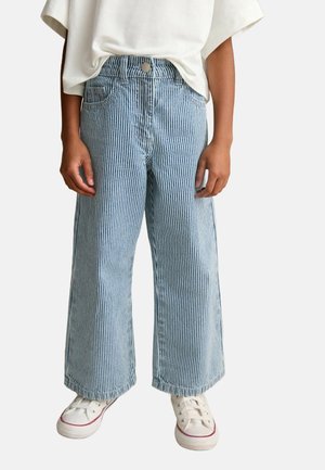 Enfant portant un pantalon en jean à jambes larges rayé bleu et blanc, des baskets blanches et un t-shirt blanc ample, debout les mains relâchées.