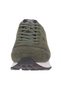 Olivgrön mockaskor med texturerad ovandel, platta skosnören och en svart tunga med vit text. Gummisula med ett distinkt mönster.